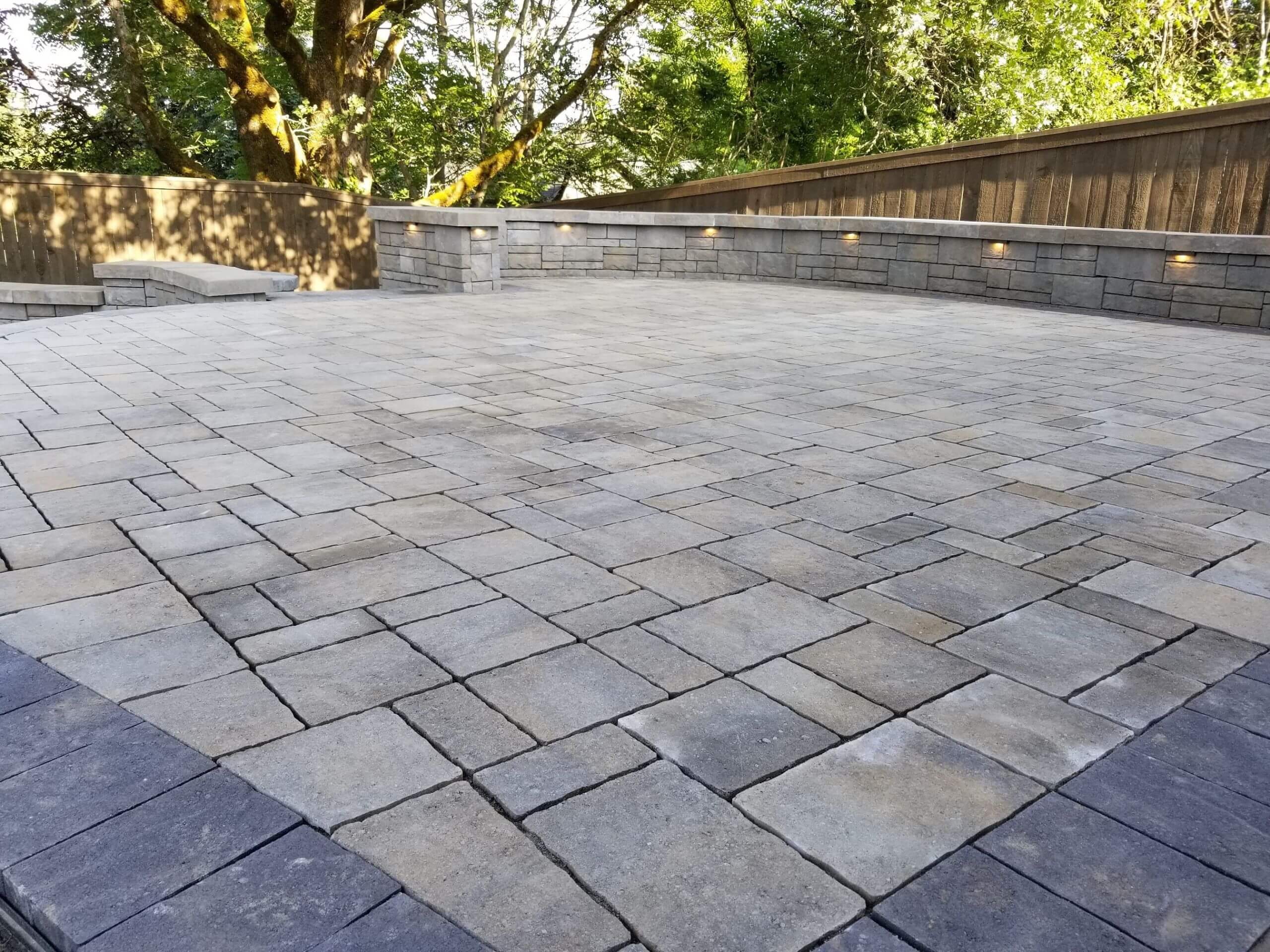 Gray paver patio bordered by a low seating wall beneath mature trees.