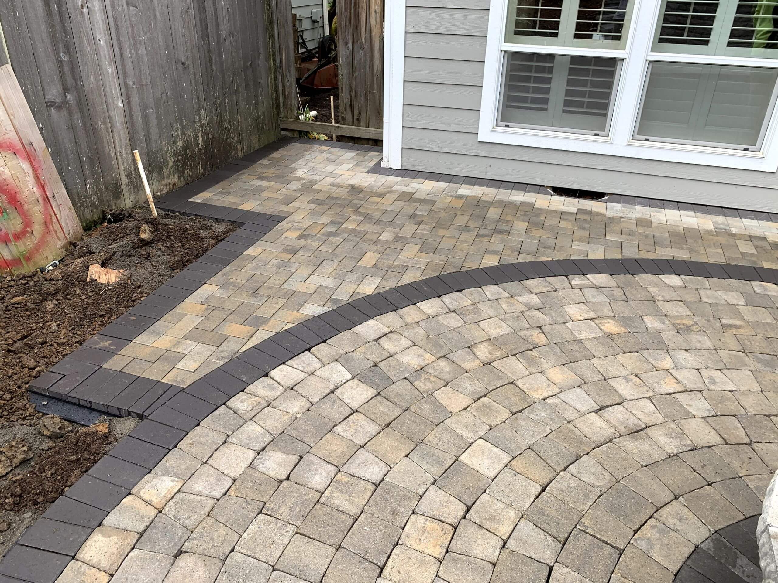 Curved dark paver border framing a tan and gray paver patio beside a house.
