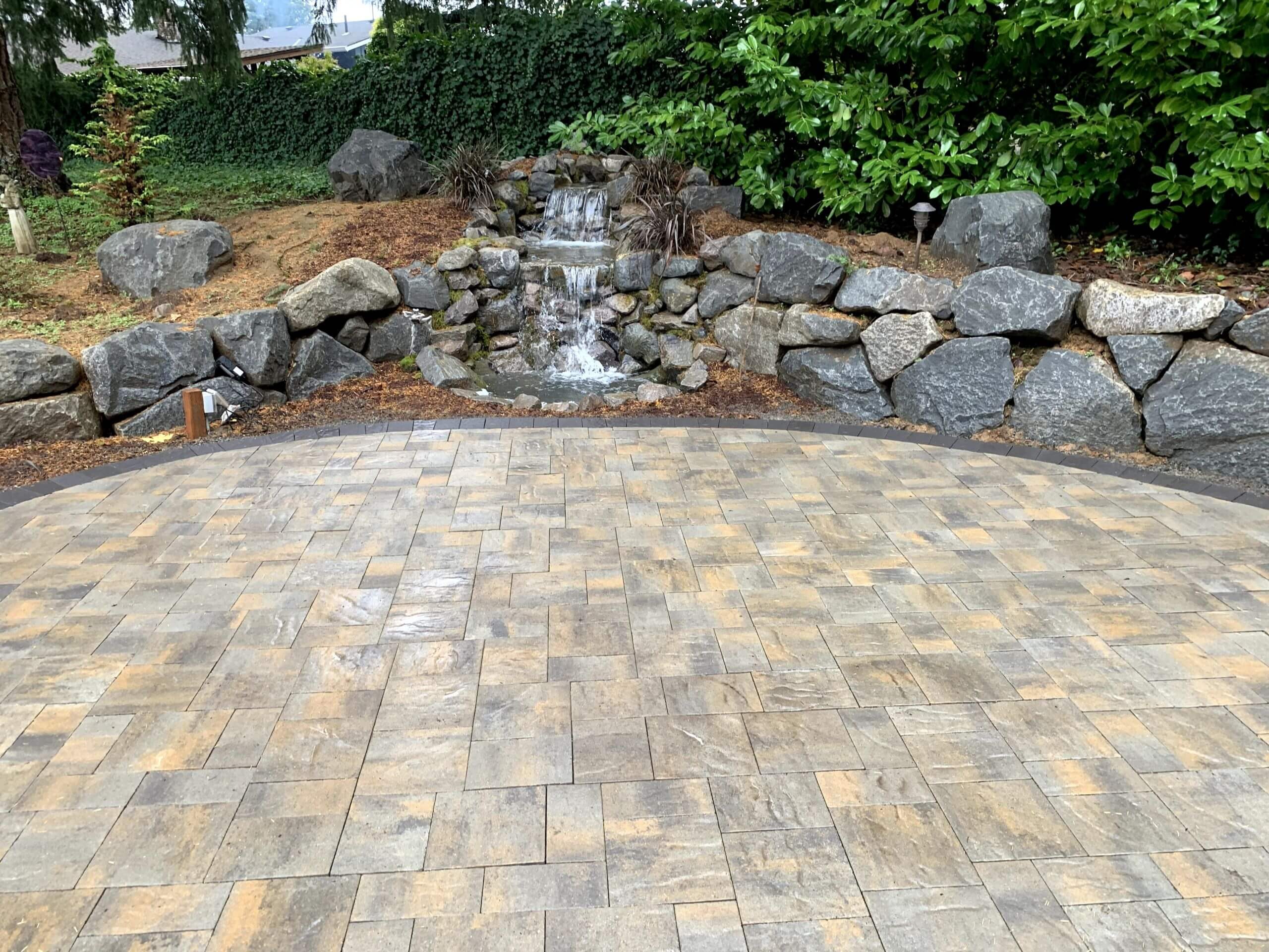 Tan and gray paver patio with a dark curved border beside a rock waterfall.