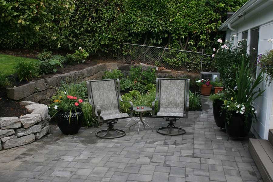 Two chairs and a small table on a gray paver patio surrounded by garden planting.